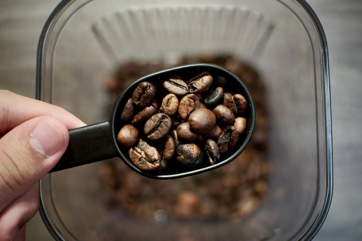 A hand holds a black scoop filled with shiny brown coffee beans over a container. The focus on the beans conveys a fresh, rich aroma.