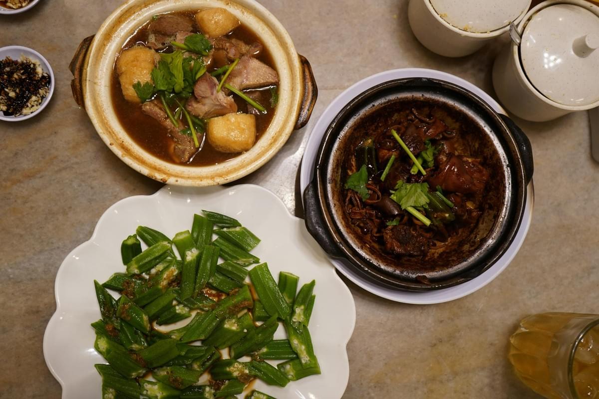 This image features a top-down view of a marble tabletop spread with clay pot dishes, sliced okra, and various condiments. The hearty meal includes a light herbal broth with meat and tofu puffs alongside a darker, braised clay pot dish topped with fresh cilantro.