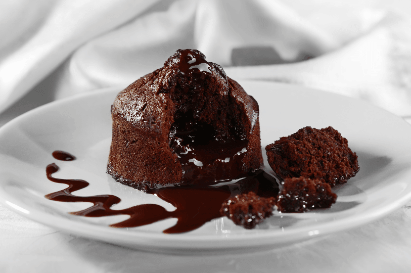 A dark chocolate lava cake with a broken center revealing a thick, slow-moving molten core. The cake is plated simply on a white dish with a decorative chocolate drizzle and crumbled cake pieces, set against a soft white background.