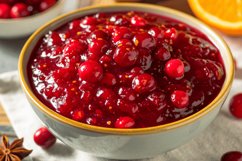 A warm, inviting bowl of homemade chunky cranberry sauce garnished with orange zest, served in a ceramic bowl alongside an orange slice and star anise on a rustic tablecloth.