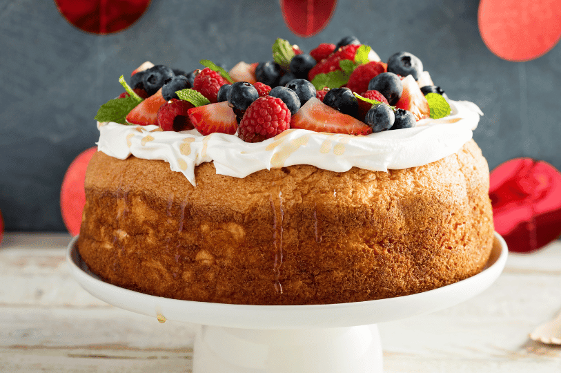 A golden sponge cake topped with whipped cream, fresh berries, and mint leaves sits on a white pedestal amid festive red accents—celebrating indulgence with vibrant color and rustic charm.