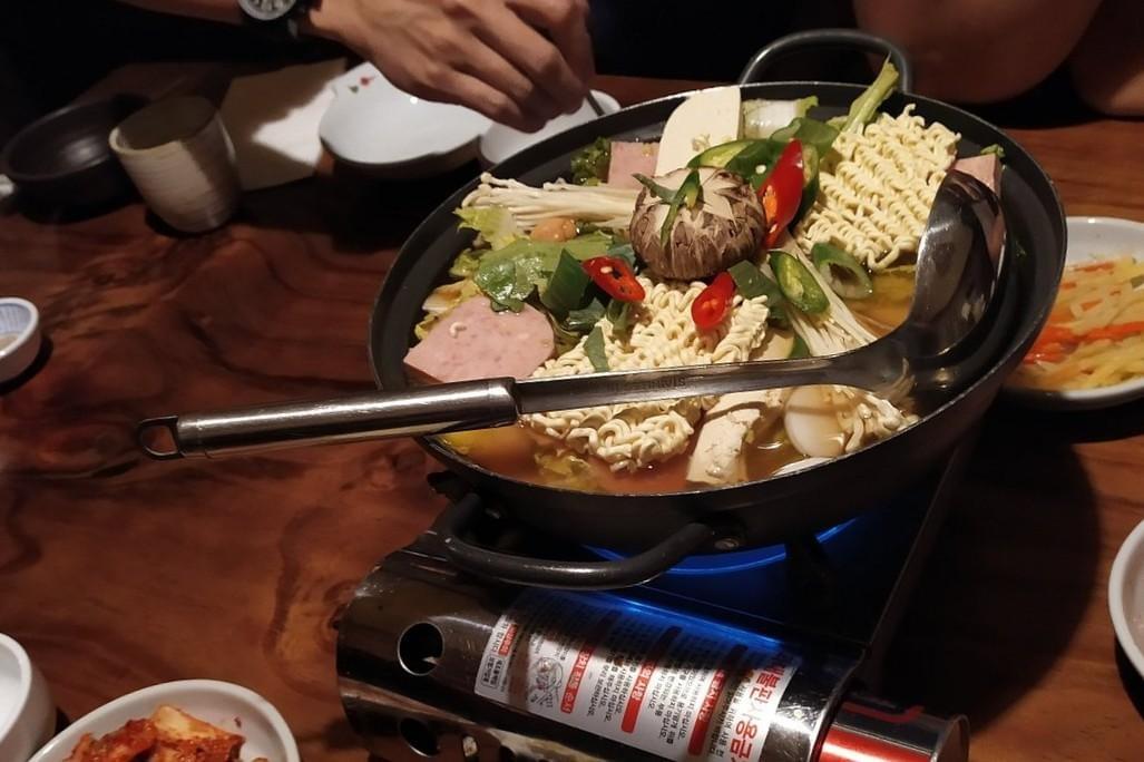An overhead shot of a large pot of Korean army stew (budae jjigae) simmering on a portable gas stove, filled with instant ramen noodles, spam, tofu, mushrooms, and vegetables. A metal ladle rests across the top of the pot, while hands and various side dishes are visible around the wooden table.