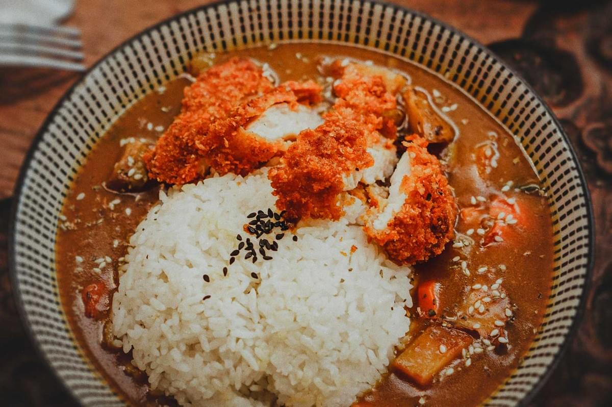 Japanese curry rice topped with crispy katsu pieces, served in a patterned bowl with vegetables and sesame seeds.