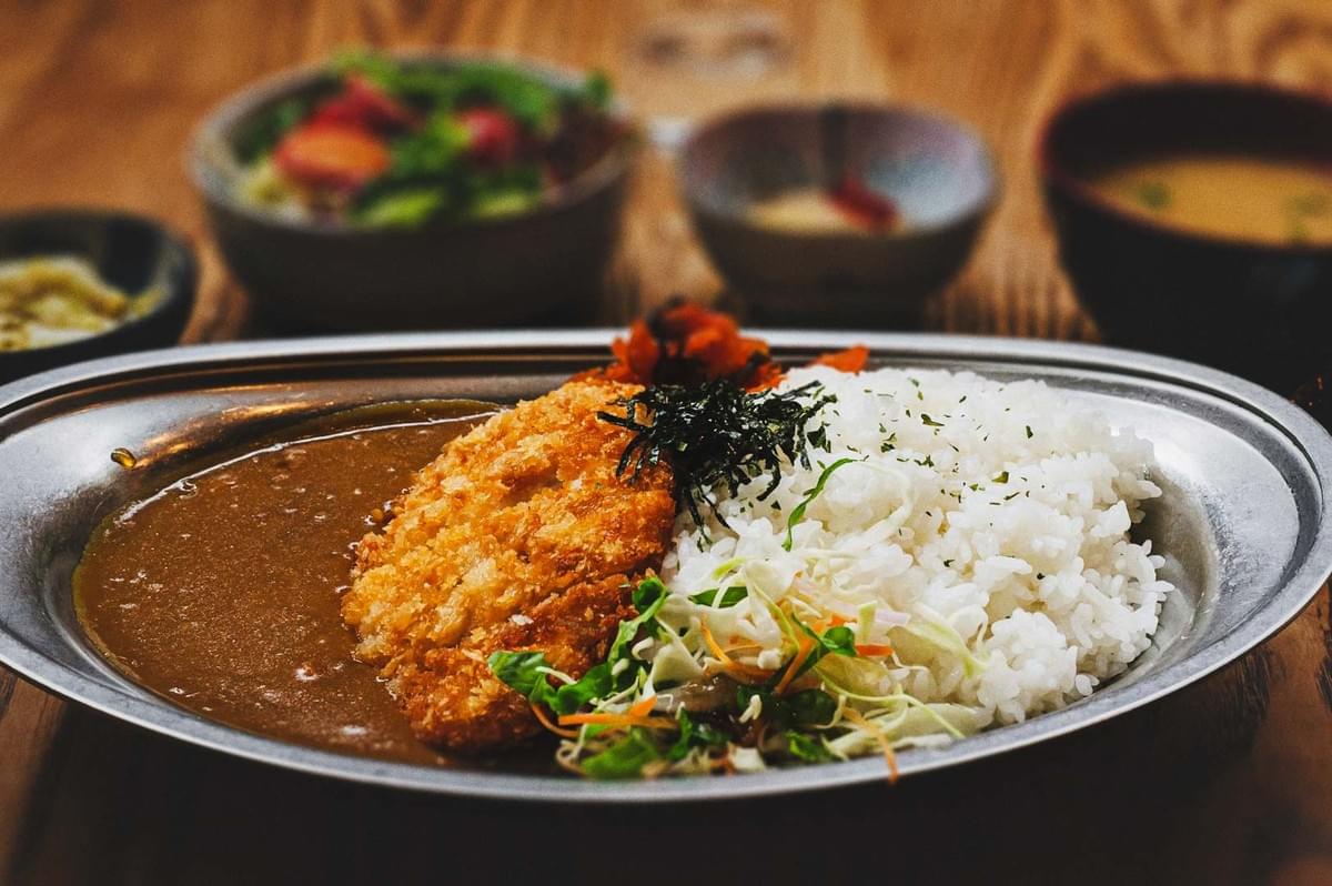 Japanese curry rice with crispy katsu, shredded cabbage, and pickles served on a metal plate with side dishes in the background.
