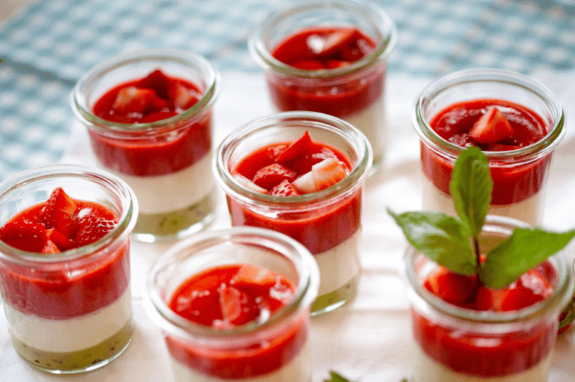 Several small glass jars filled with layers of creamy Panna Cotta topped with a thick strawberry sauce and fresh sliced strawberry pieces.