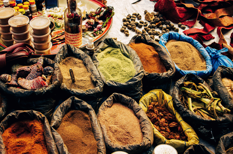 A bustling market stall display with large open sacks filled with various ground spices in shades of ochre, sage green, and deep brown. Nearby, a metal tray holds small glass vials and bottles of traditional oils and essences.