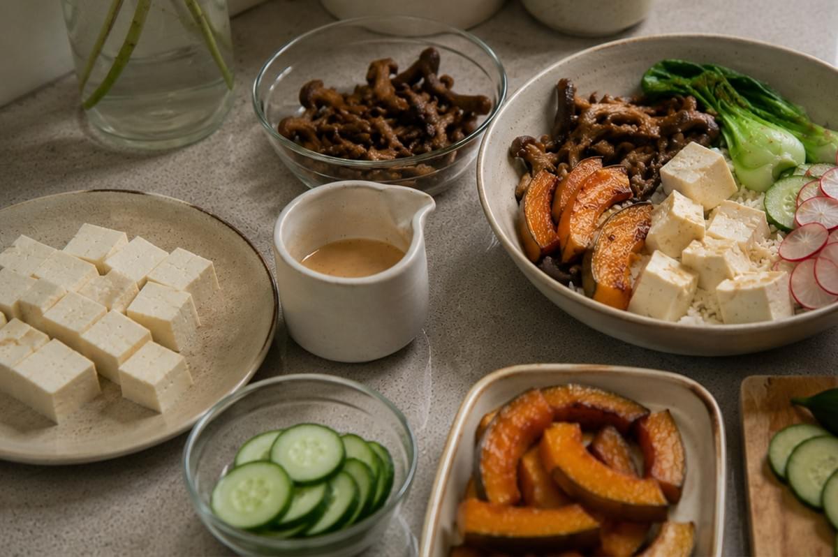 A high-angle, eye-level shot showcasing the various components of a vegetarian bowl laid out on a kitchen counter. A large bowl to the right is already filled with rice, tofu, roasted pumpkin, mushrooms, bok choy, radishes, and cucumbers. Surrounding the bowl are small glass and ceramic dishes containing the individual ingredients, including a plate of raw tofu cubes, a bowl of cooked mushrooms, a bowl of roasted pumpkin slices, a small bowl of sliced cucumbers, a small pitcher of dressing, and a glass of water, highlighting a "mise en place" kitchen scene.