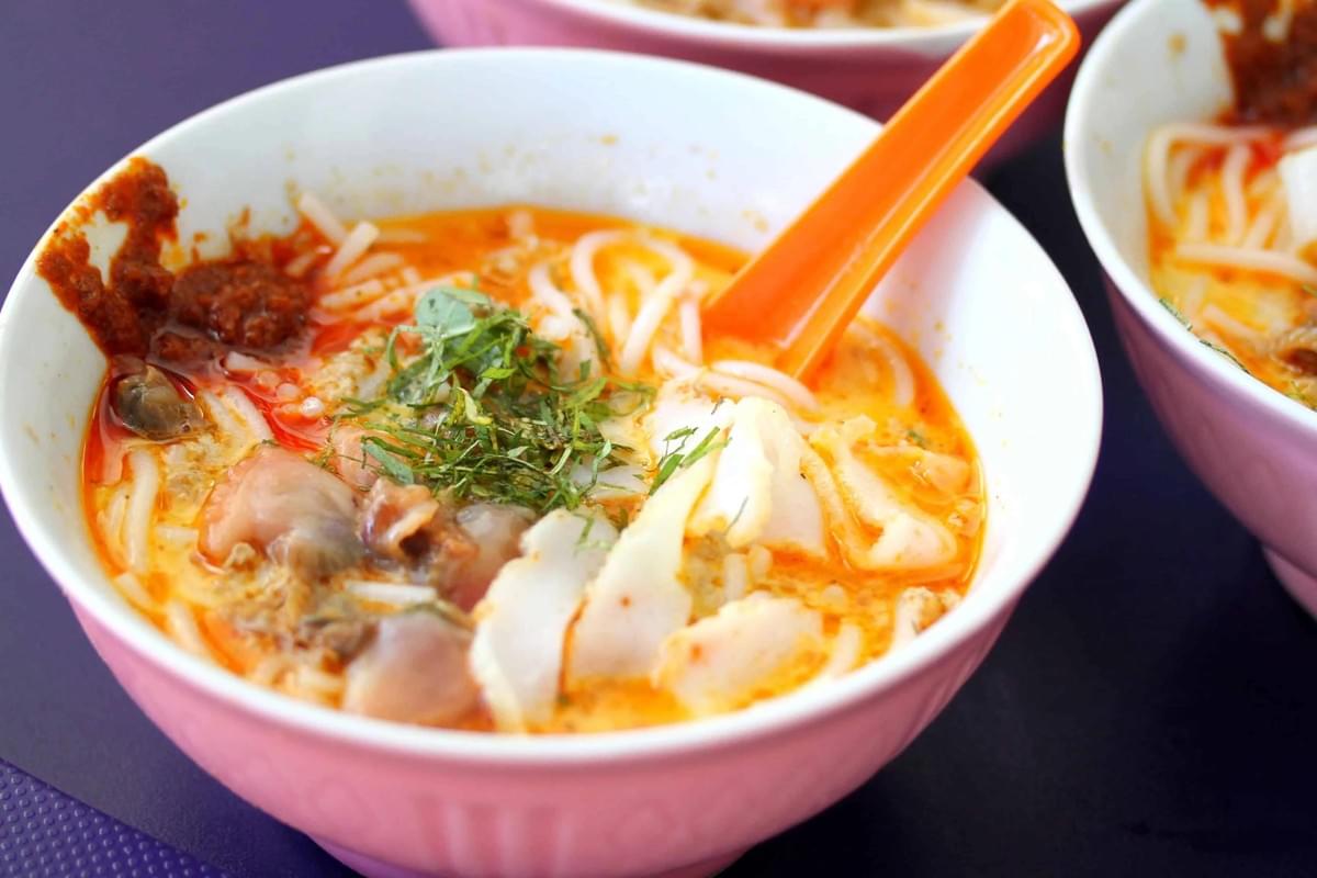 A bowl of laksa features thick rice noodles in a rich orange broth, garnished with sliced fish cakes and finely shredded herbs. A dollop of dark red sambal sits on the rim of the bowl next to an orange plastic spoon.