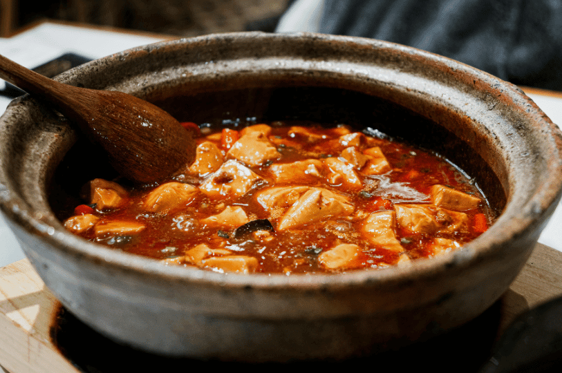 A traditional clay pot filled with simmering Mapo Tofu, featuring a deep red, oily chili sauce and large tofu cubes, with a wooden ladle resting inside.