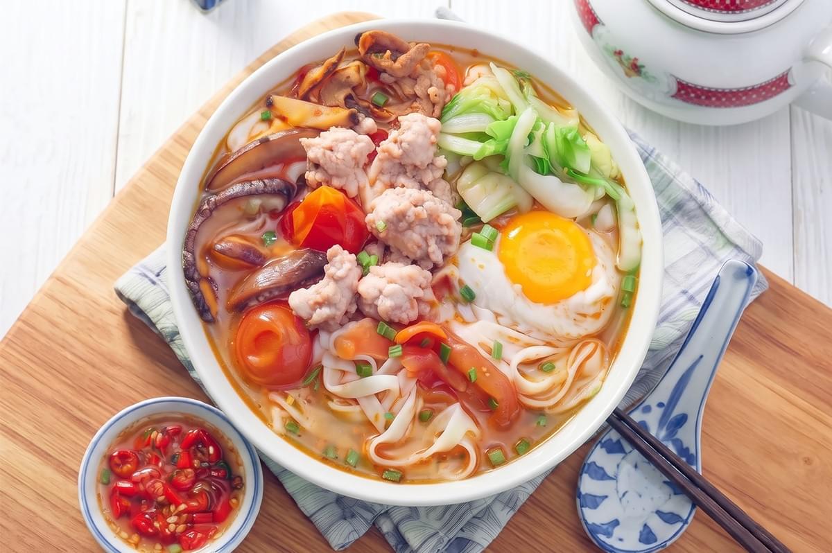 Bowl of homemade ban mian noodle soup with minced meat, poached egg, vegetables, and chili sauce on a wooden board.