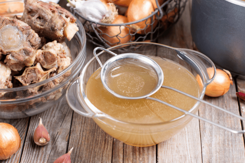 A glass bowl of freshly strained liquid gold stock next to a bowl of roasted marrow bones, garlic, and onions used for the infusion.