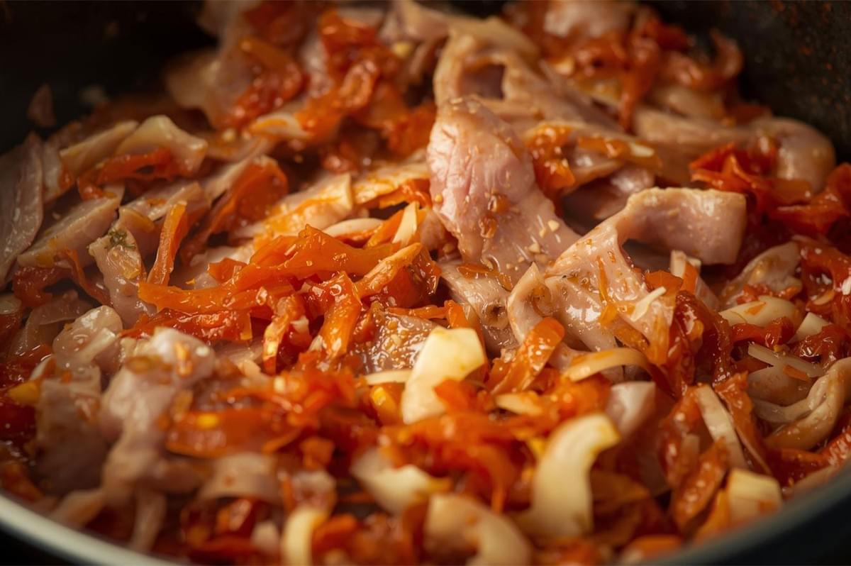 A close-up of the stew base simmering in a pan, showing raw or lightly cooked sliced pork, chopped onions, and sliced red kimchi being stir-fried before the broth is added.