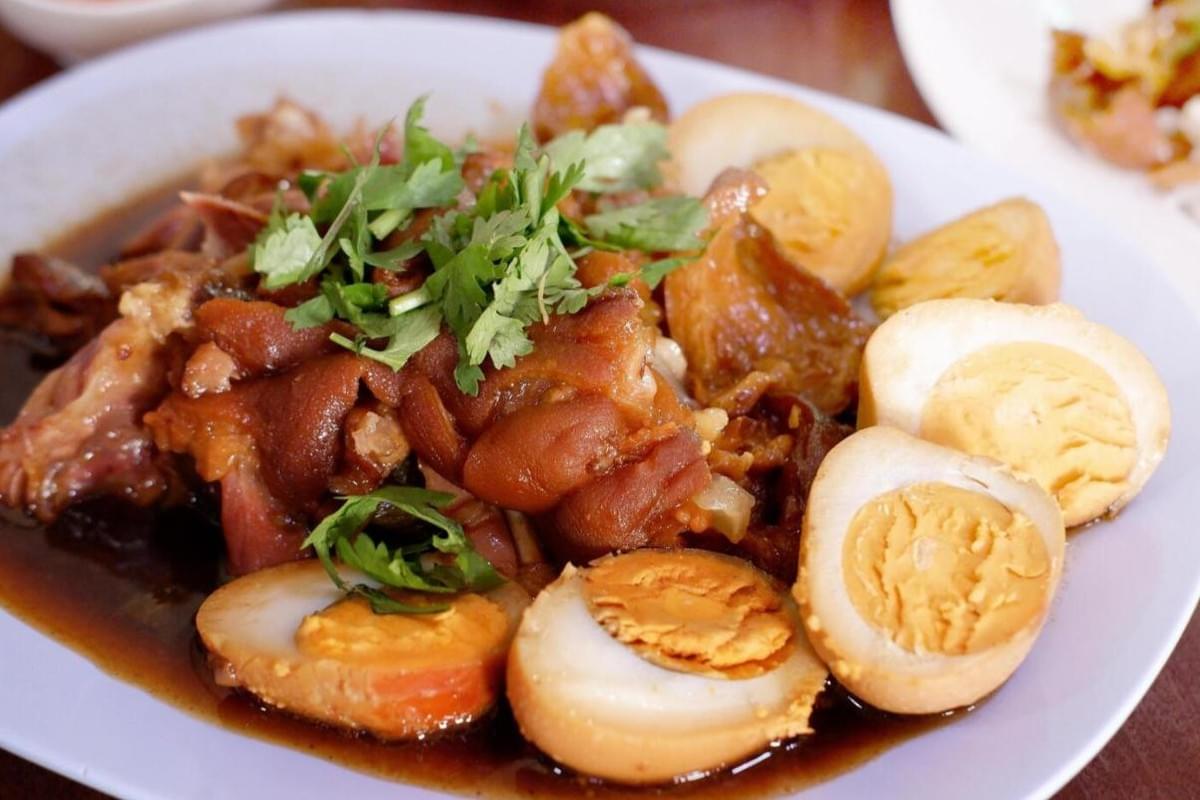  This image features a plate of Rojak, a Thai braised pork leg dish served with halved hard-boiled eggs in a rich, dark soy-based sauce. The tender pieces of pork are topped with fresh green coriander leaves and arranged on a light blue plate, highlighting the contrast between the savory meat and the bright garnish.