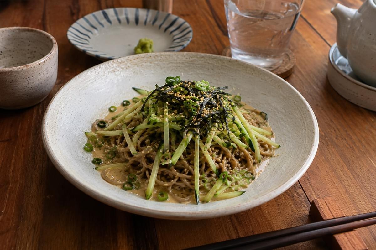A finished plate of cold soba noodles sits beautifully presented on a warm, rustic wooden table. The noodles are piled high in a large, speckled white ceramic dish, heavily garnished with julienned cucumbers, scallions, sesame seeds, and a crown of shredded nori, with the creamy sauce visible at the base of the pile. The dining setup is completed by a small empty ceramic cup, a blue-and-white striped accent plate with a small dollop of green wasabi, a glass of ice water, a white ceramic teapot, and dark wooden chopsticks resting on a small wooden holder in the foreground.