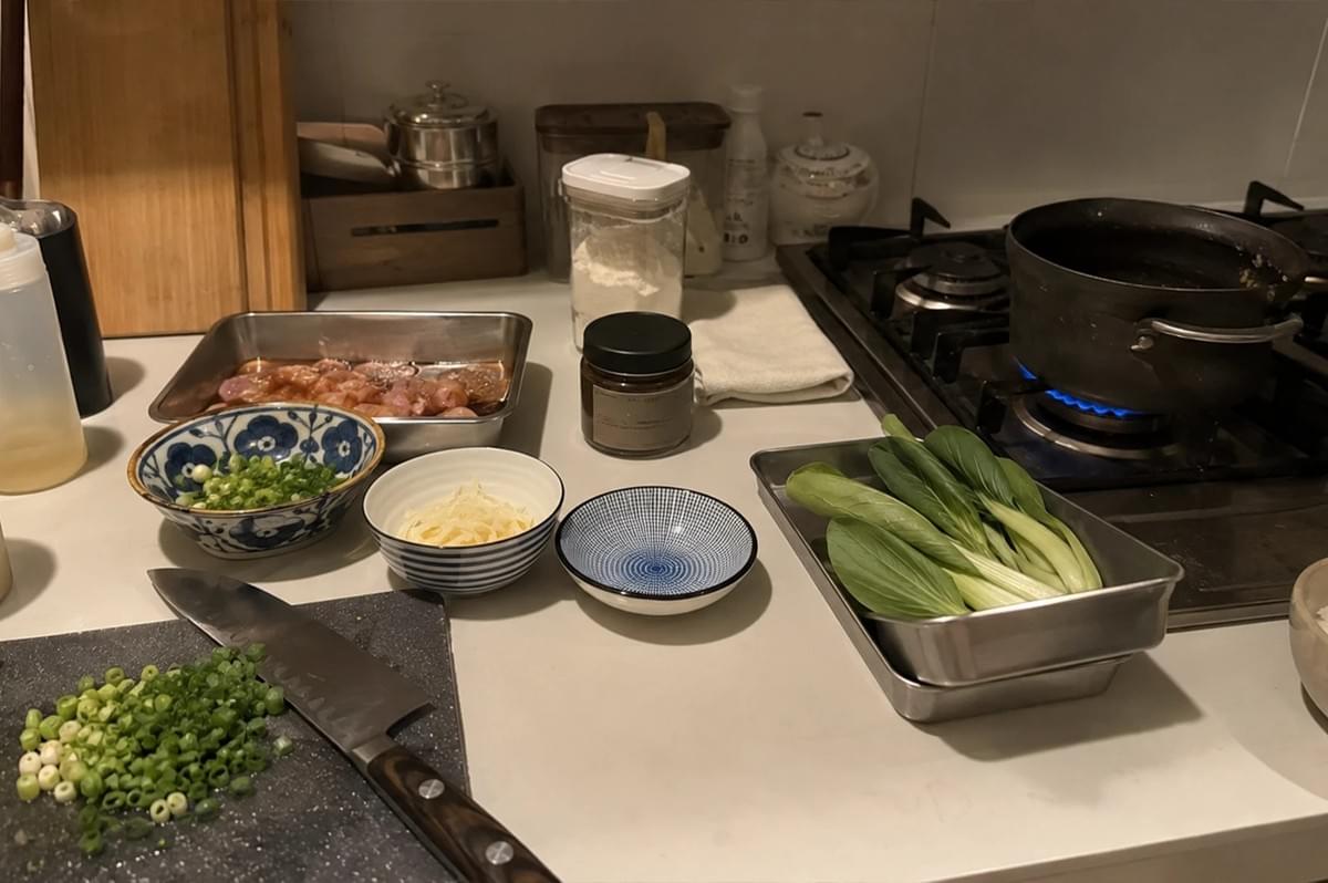 !The image captures a well-organized kitchen counter during food preparation, showcasing various fresh ingredients and cooking essentials. On a dark cutting board in the foreground sits a sharp kitchen knife next to a small pile of finely chopped scallions, while nearby, a blue-patterned bowl holds additional chopped scallions and a smaller bowl contains thin strips of ginger. A metal tray holds marinated pieces of meat, another tray is filled with fresh bok choy, and a small bowl of cooked white rice sits near the edge of the counter. In the background, a dark pot rests on a lit gas stove burner, and various jars, containers, and a wooden block are neatly arranged against the wall, contributing to the busy yet functional atmosphere of a home kitchen in the middle of cooking a meal.  The image captures a well-organized kitchen counter during food preparation, showcasing various fresh ingredients and cooking essentials. On a dark cutting board in the foreground sits a sharp kitchen knife next to a small pile of finely chopped scallions, while nearby, a blue-patterned bowl holds additional chopped scallions and a smaller bowl contains thin strips of ginger. A metal tray holds marinated pieces of meat, another tray is filled with fresh bok choy, and a small bowl of cooked white rice sits near the edge of the counter. In the background, a dark pot rests on a lit gas stove burner, and various jars, containers, and a wooden block are neatly arranged against the wall, contributing to the busy yet functional atmosphere of a home kitchen in the middle of cooking a meal.