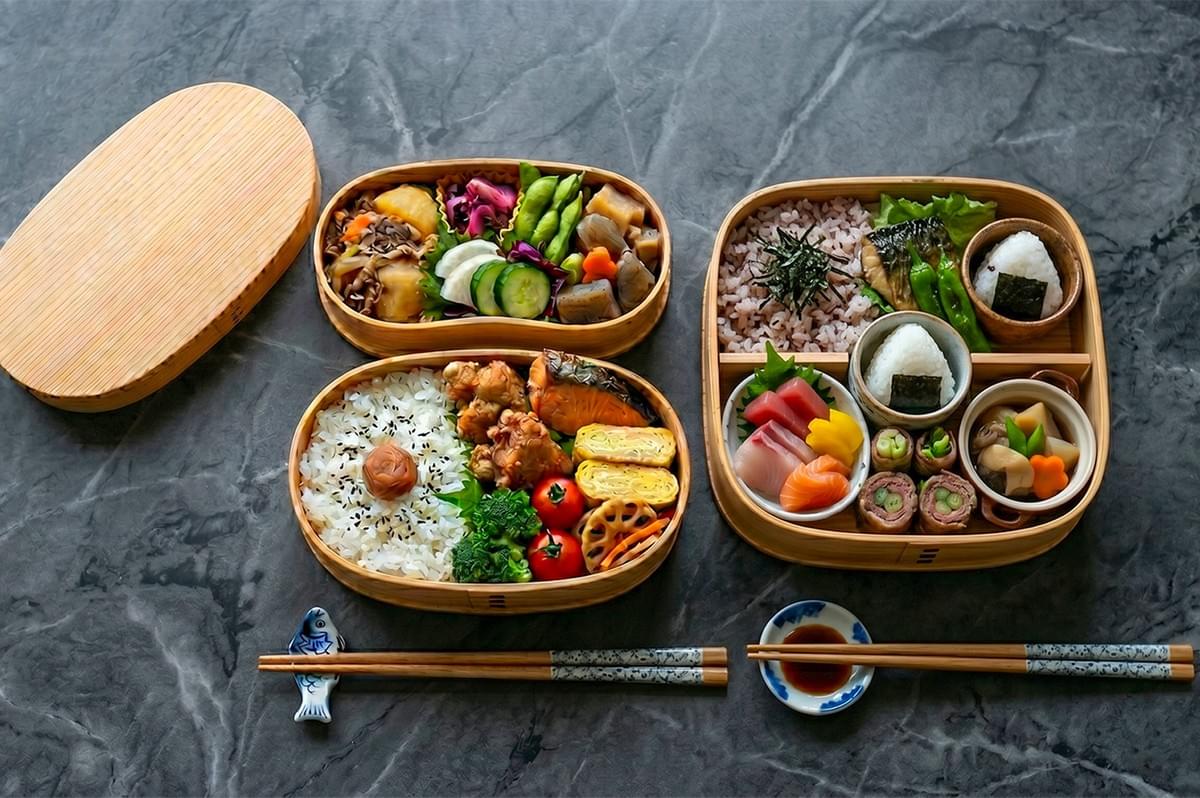 Traditional Japanese bento boxes filled with rice, sashimi, tamagoyaki, grilled salmon, and assorted vegetables on a grey marble tabletop.