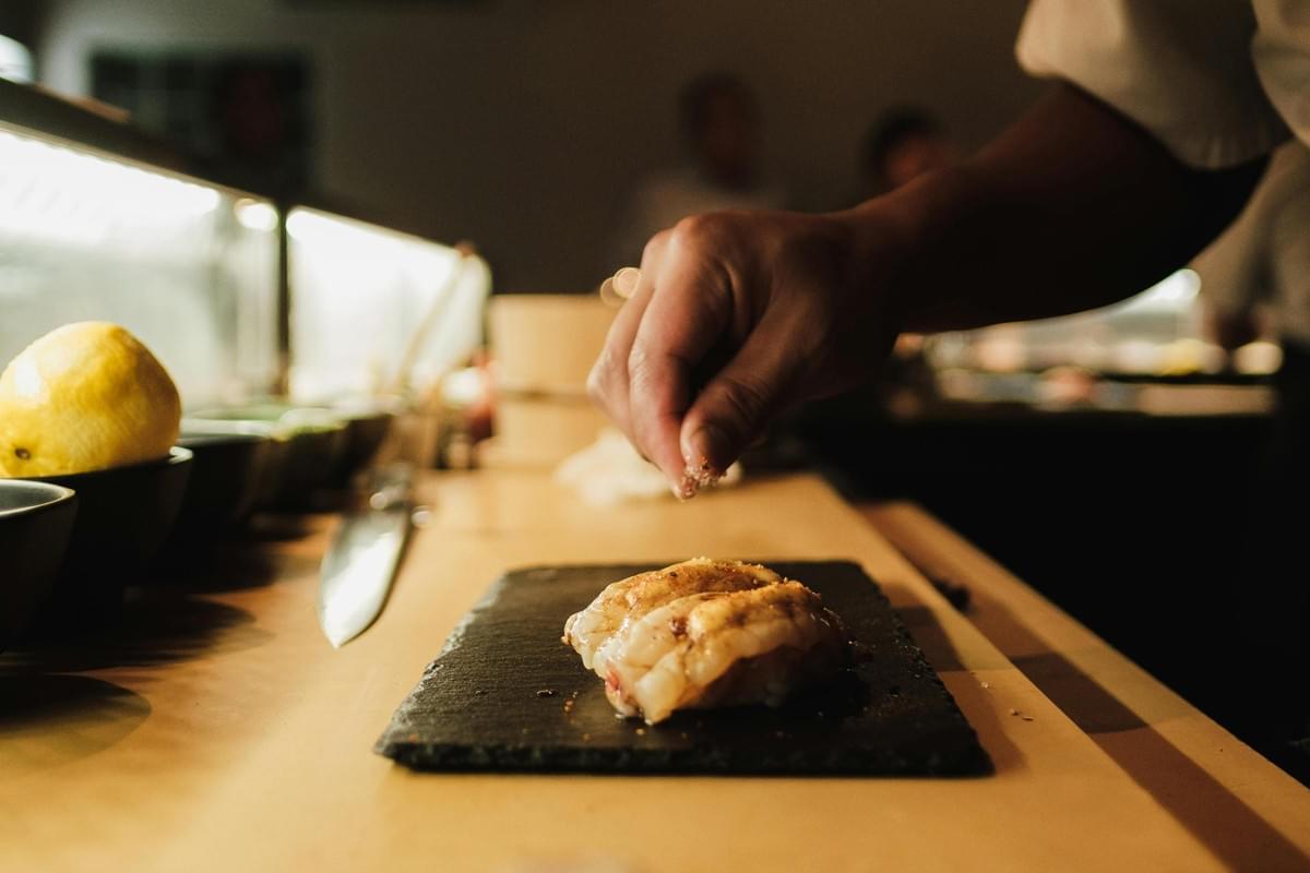 These close-up images showcase the precise culinary artistry of a chef, capturing hands in the process of delicately garnishing and seasoning sushi on a black slate platter. The warm, focused lighting accentuates the textures of the fresh seafood and the wooden countertop, emphasizing the attention to detail involved in the preparation.