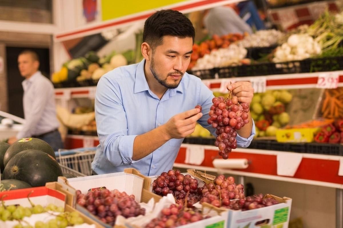 Man in a light blue shirt examining a bunch of red grapes in the produce section of a grocery store