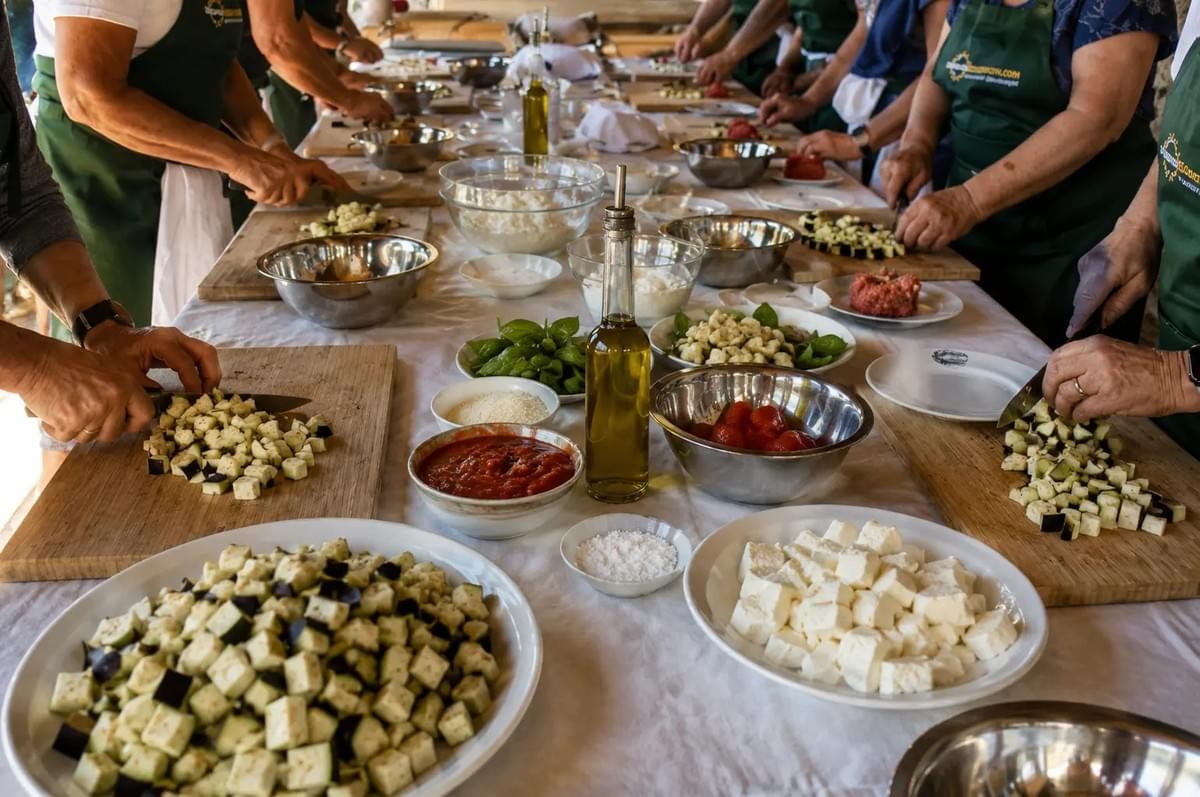 A multi-generational family enjoys a festive outdoor Greek Easter feast at a sunlit rustic courtyard table laden with traditional foods. The family members are tapping dyed red eggs together in a celebratory toast, with a large braided Tsoureki Easter bread featured in the foreground.