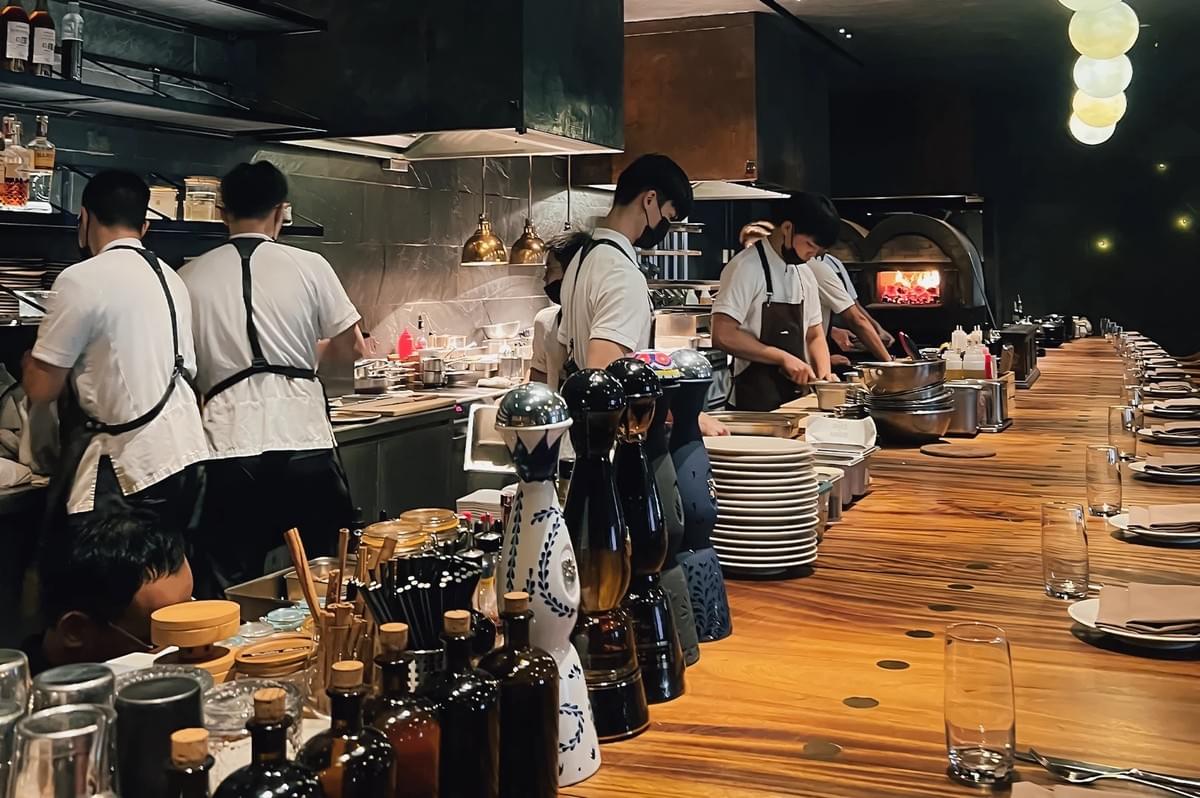 Wide-angle interior shot of the Burnt Ends open kitchen in Singapore, showing chefs working behind a long wooden counter with fire-driven grills, highlighting the restaurant’s energetic fine dining atmosphere and iconic romantic dining experience
