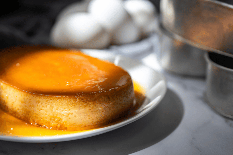 A wide, oval-shaped flan resting on a white plate with visible air bubbles in the custard, set against a background of fresh eggs and metal baking tins.