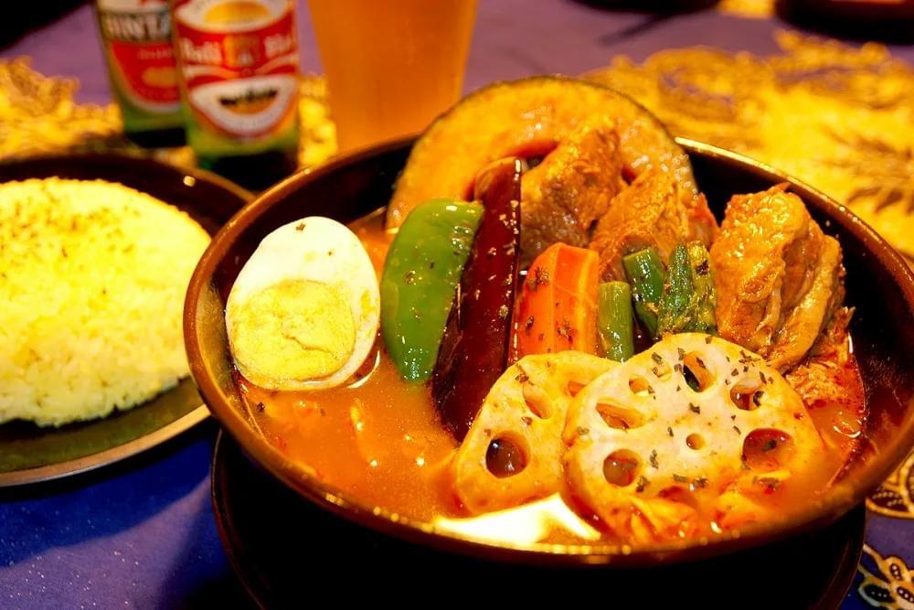 A bowl of rich, orange-tinted Japanese soup curry is filled with chunks of meat, a hard-boiled egg half, and vibrant vegetables like lotus root, green pepper, and eggplant. In the background, a side plate of yellow turmeric rice and a tall glass of beer sit on a patterned blue tablecloth.