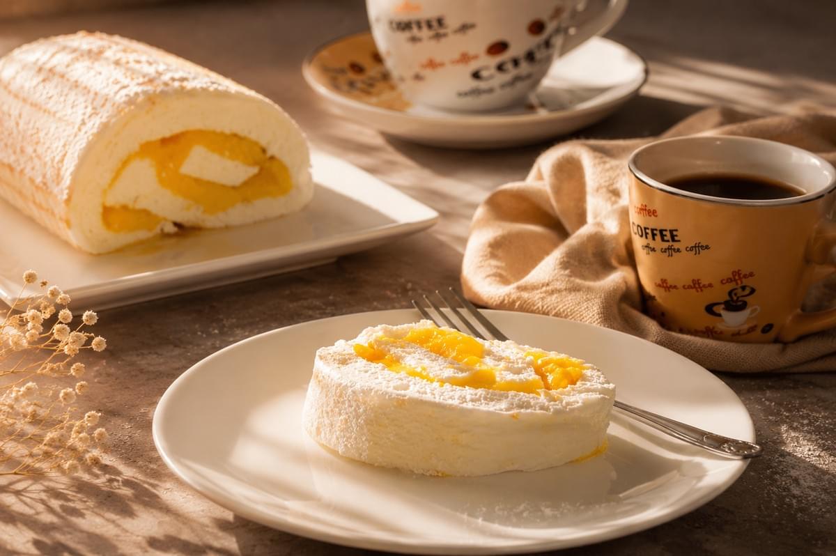 A slice of meringue roll cake with yellow custard filling served on a small plate alongside a cup of coffee and the remaining cake roll in the background.