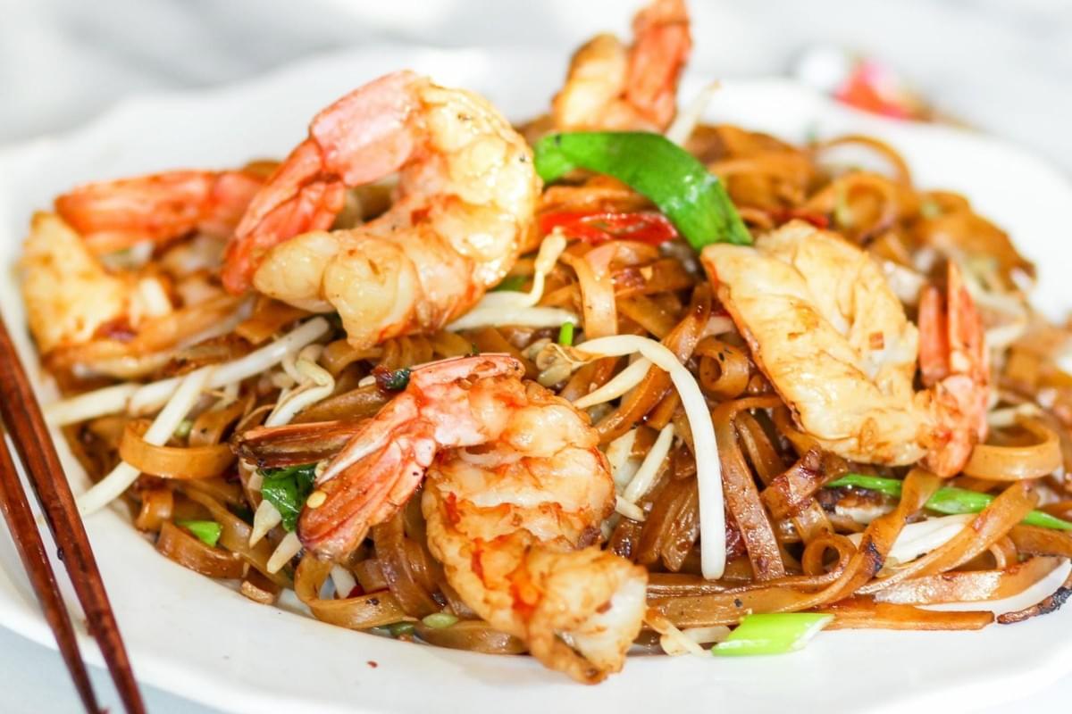 A plate of stir-fried flat rice noodles is tossed with bean sprouts and green onions, served with a pair of chopsticks on the side. The dish is topped with several large, succulent prawns that have been cooked to a bright orange hue.