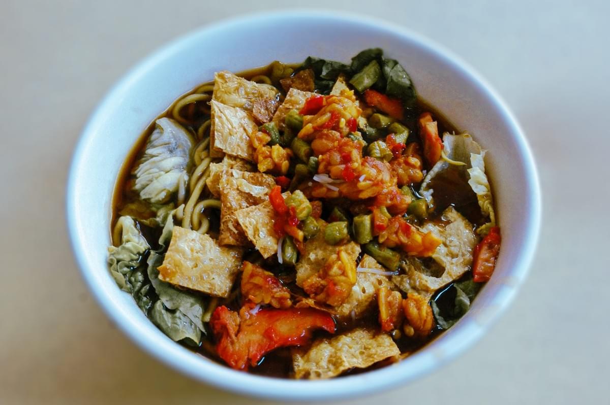 Bowl of vegetarian noodles in broth topped with tofu skin, chopped pickled vegetables, leafy greens, and sliced mock meat.
