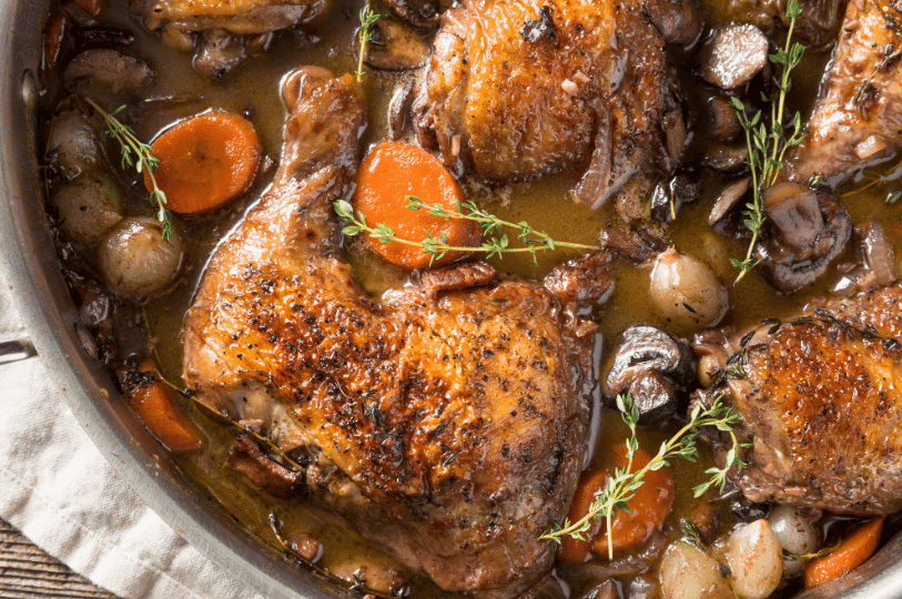A detailed macro shot of Coq au Vin showing the rich texture of seared chicken skin, pearl onions, and carrots in a wine reduction.