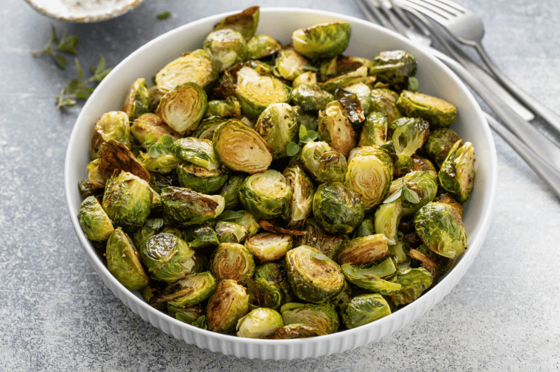 A close-up of perfectly charred and golden-brown roasted Brussels sprouts in a white ceramic bowl, highlighted by natural side lighting.
