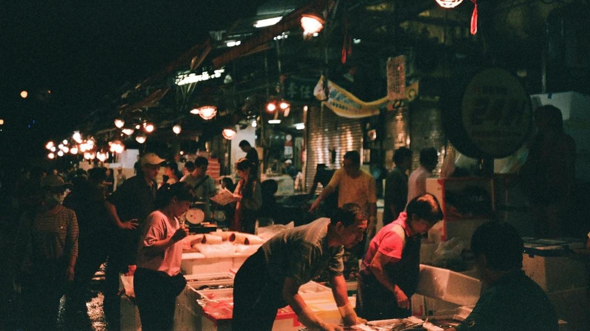 A bustling night market scene is captured in low light, featuring vendors and customers interacting over rows of styrofoam crates likely containing fresh seafood or produce. Warm overhead string lights illuminate the crowd and reflect off the wet pavement, creating a vibrant and authentic atmospheric glow.