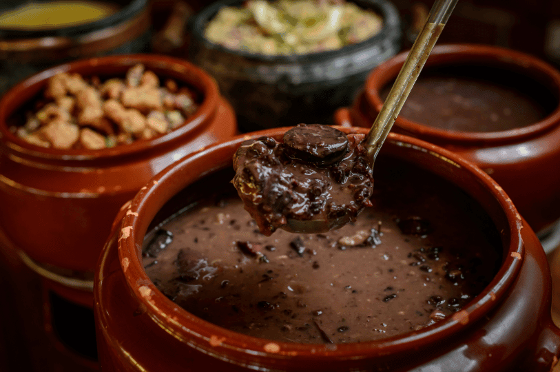 A rustic clay pot of Brazilian feijoada—rich with black beans and pork—sits center stage, with a ladle lifting a hearty portion and side dishes in surrounding pots, evoking the warmth of traditional communal dining.