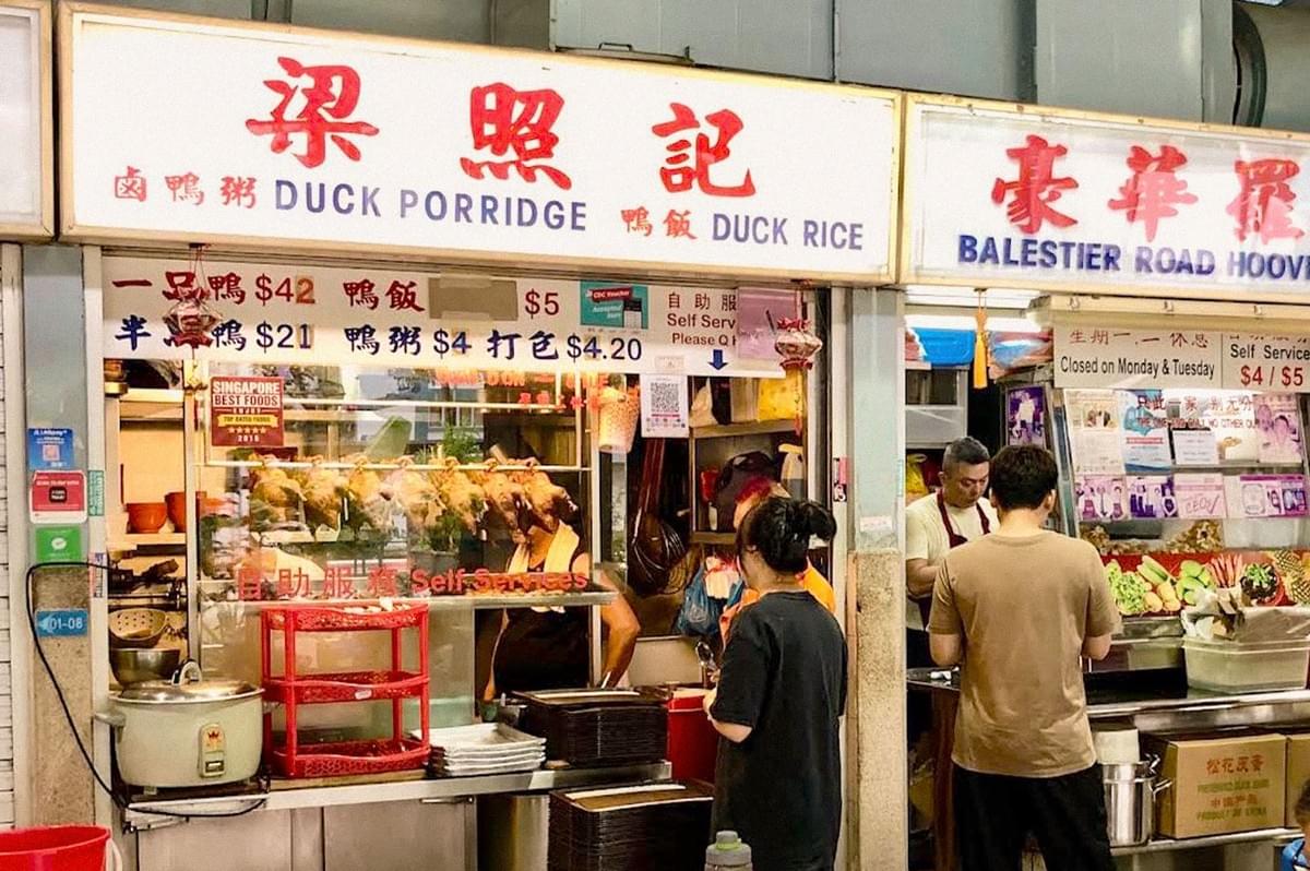 A Singapore hawker stall serving duck porridge and duck rice, with roasted ducks displayed behind the counter and customers ordering food.