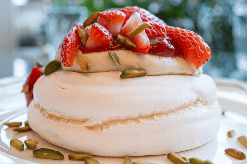 A close-up of a large, circular pavlova showing a smooth, crisp meringue base topped with thick cream, strawberry halves, red fruit coulis, and toasted pistachios.
