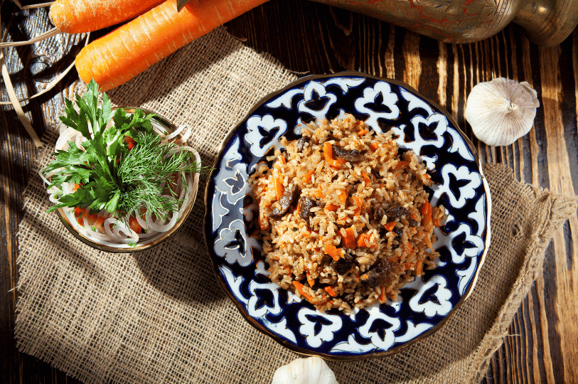 Overhead view of traditional Uzbek plov on a blue patterned plate, featuring rice, carrots, and meat, served alongside an onion and herb side salad on a rustic burlap surface.