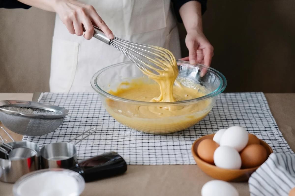 A person whisking a thick, golden batter in a glass bowl on a checkered cloth, surrounded by eggs and measuring tools.