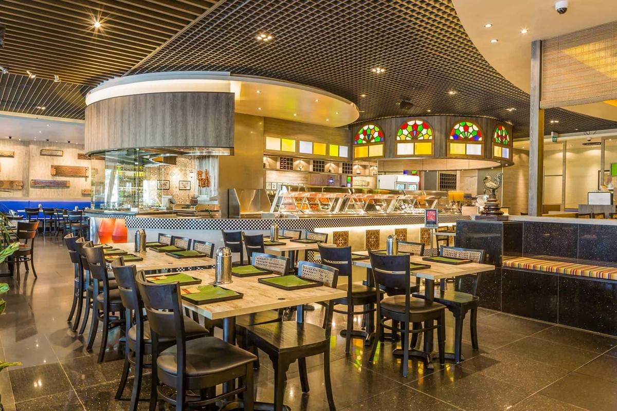 The image shows the brightly lit, modern interior of a restaurant with neatly arranged dining tables set with green placemats and black chairs. In the background, a curved buffet station and an open kitchen area are visible beneath a textured, dark ceiling accented by colorful stained-glass features.