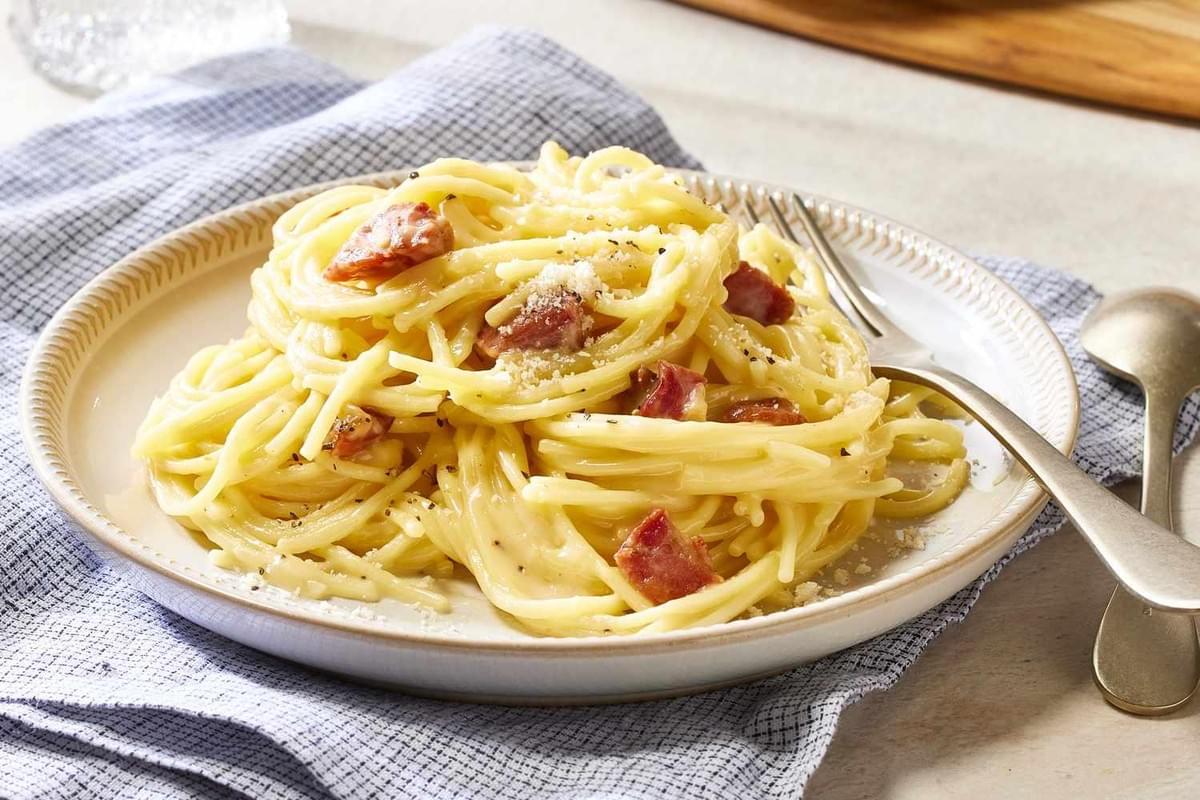 A plate of creamy spaghetti carbonara is piled high and topped with pieces of crispy, browned meat and grated cheese. The dish is sprinkled with freshly cracked black pepper and sits on a beige plate next to a silver fork and spoon.