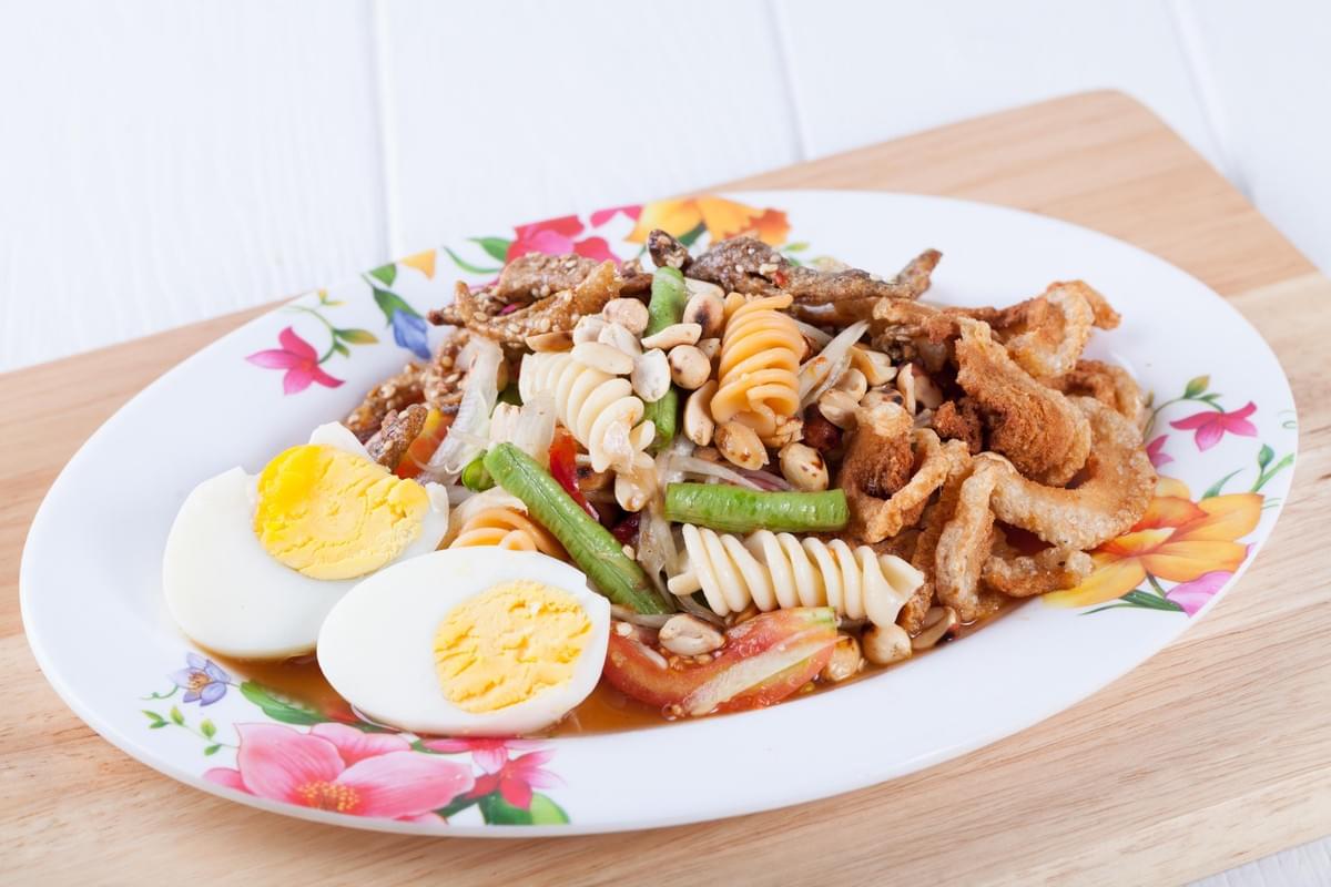 This image showcases a unique take on Rojak, a Thai papaya salad mixed with fusilli pasta, long beans, peanuts, and tomatoes. The dish is presented on a floral-patterned oval plate, complete with hard-boiled egg halves, crispy pork cracklings, and small fried fish for a variety of textures.