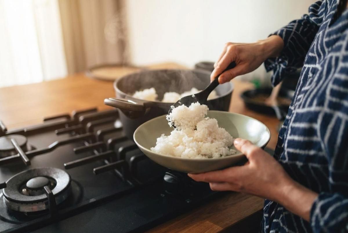 Person scooping freshly steamed white rice from a cooking pot into a shallow bowl.