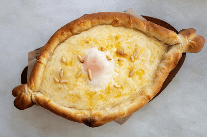 An overhead shot of a Khachapuri with a unique twist: the cheese filling is topped with a lightly poached egg and sprinkled with toasted pine nuts. The bread boat features decorative knotted ends and sits on a wooden serving paddle.