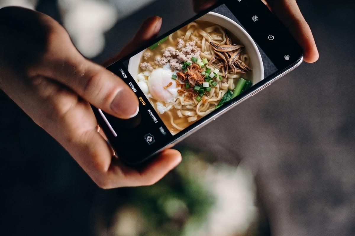 Person's hands holding a smartphone to take a photo of a bowl of ban mian noodle soup.