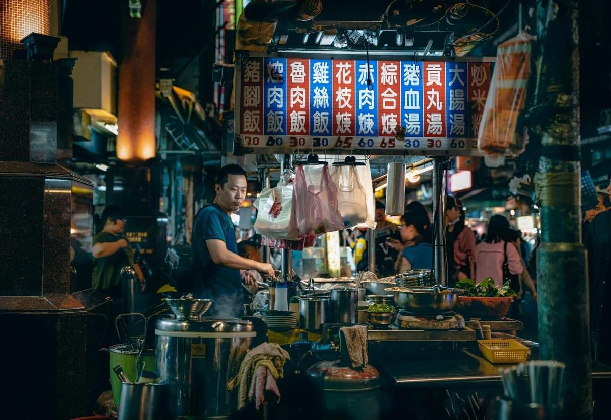 Night market food stall vendor cooking under a neon menu sign with Chinese characters.