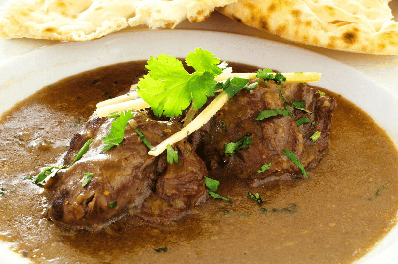 Tender chunks of slow-cooked beef Nihari, garnished with ginger julienne and fresh cilantro, with naan bread visible in the background.