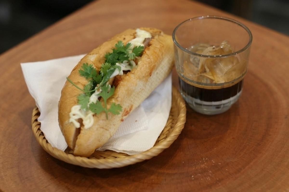 Banh mi sandwich topped with fresh cilantro and mayo on a woven tray, paired with iced Vietnamese coffee on a wooden table.
