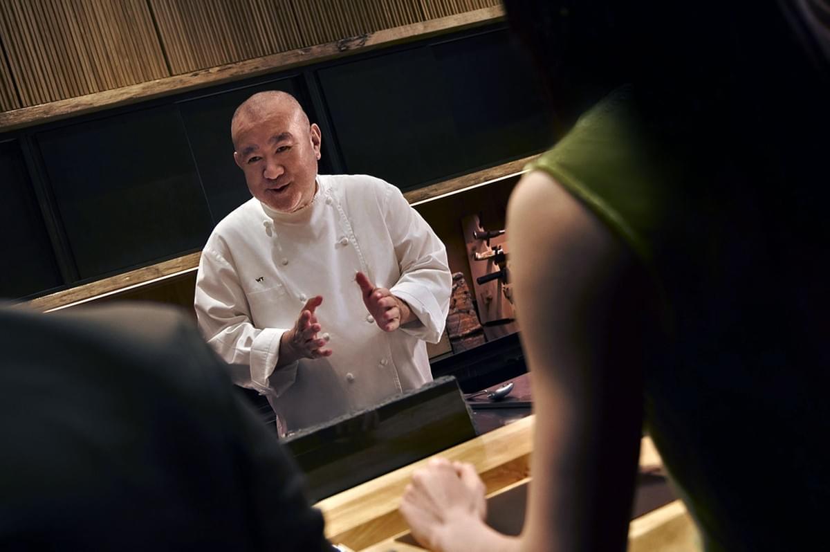 A chef in a white uniform, smiling warmly, engages with two diners seated at a wooden counter. The setting is cozy and intimate, with a focus on culinary craft.