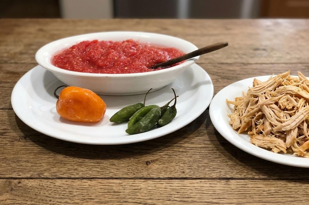 Traditional Bolivian llajua salsa in white bowl with fresh orange and green chili peppers, served alongside shredded chicken on wooden table