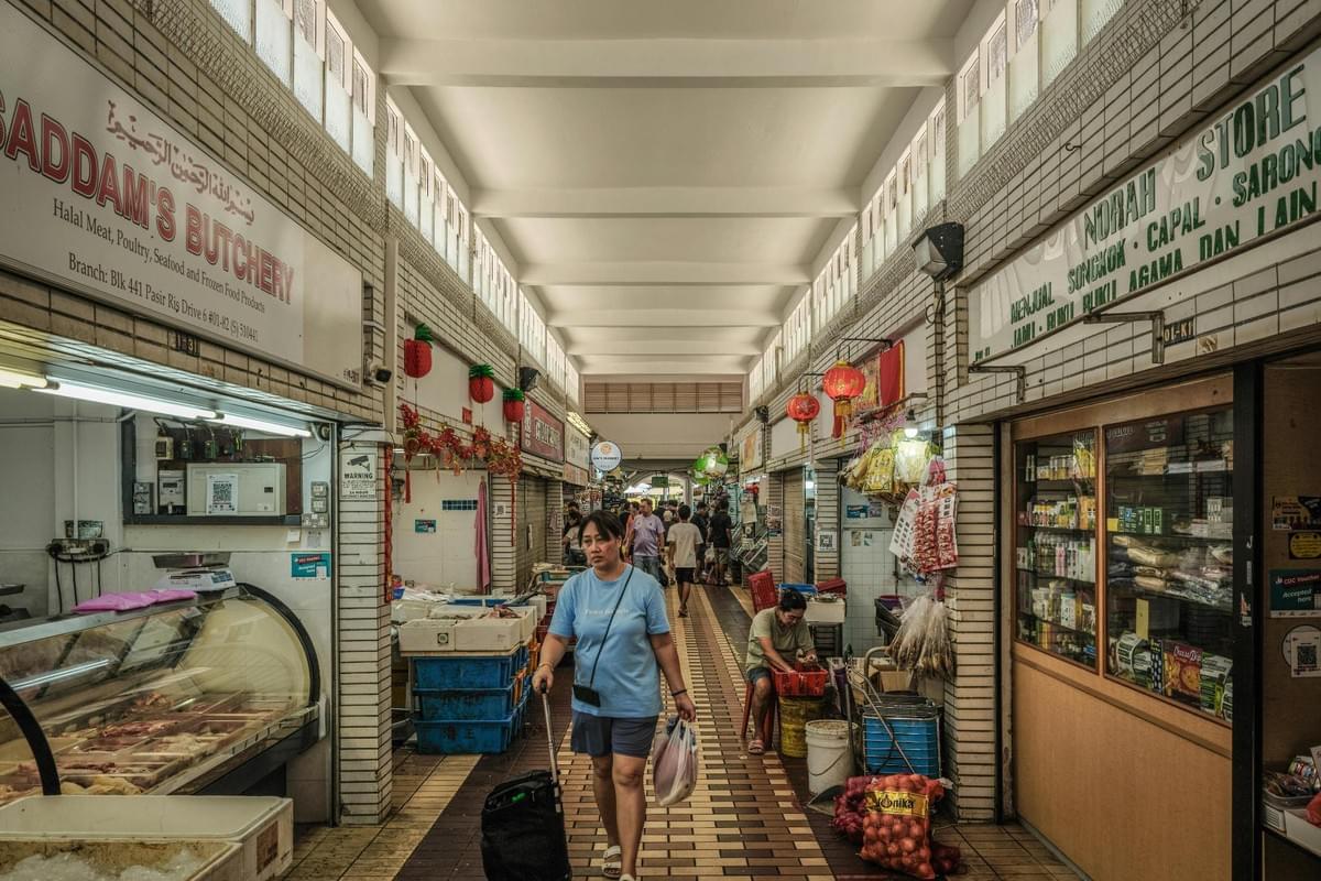 The image depicts a bustling corridor in a Singaporean market, featuring a butchery and a general store adorned with traditional red lanterns. A woman in a blue shirt walks toward the foreground with a shopping trolley while vendors and other shoppers populate the background of the narrow, tile-lined walkway.