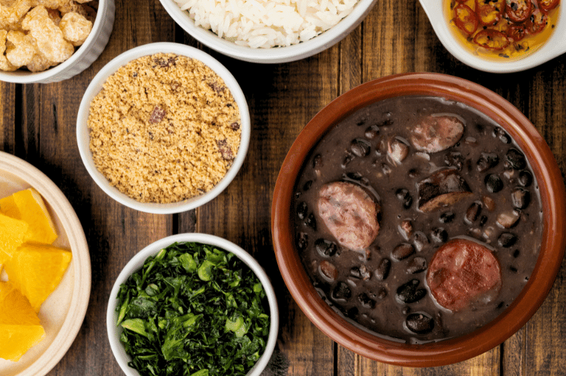 A bowl of Brazilian feijoada—black beans simmered with pork and sausage—anchors a rustic spread of rice, collard greens, farofa, orange slices, pork rinds, and chili oil, celebrating the dish’s bold flavors and festive tradition.